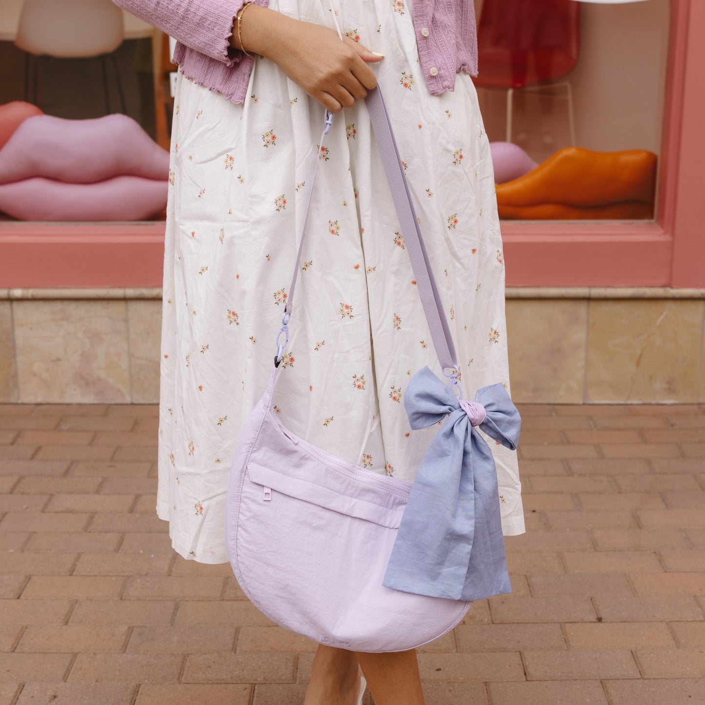 Woman in a white dress with a purple cardigan and matching bag standing on a brick pavement.