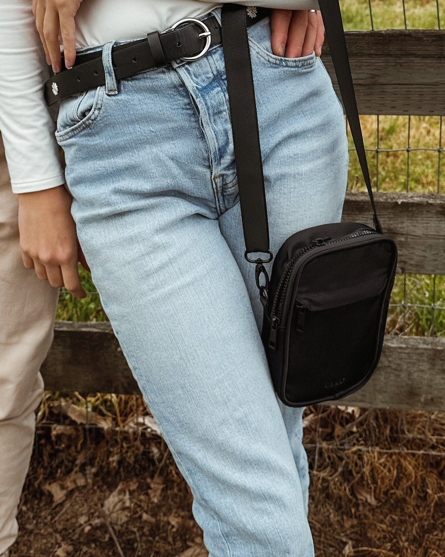 Person wearing a white shirt and light blue jeans with a black crossbody bag in an outdoor setting.